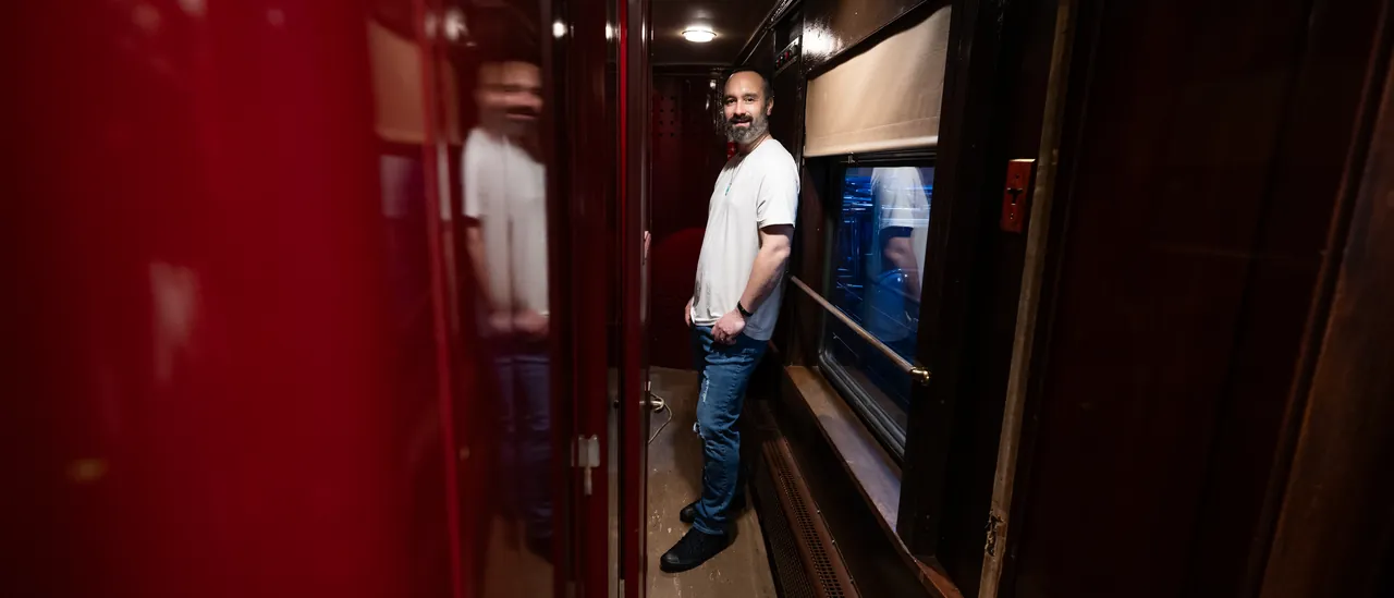 Michael Streck standing in the dim, wood-panelled corridor of a vintage railway carriage, beside a lacquered red door